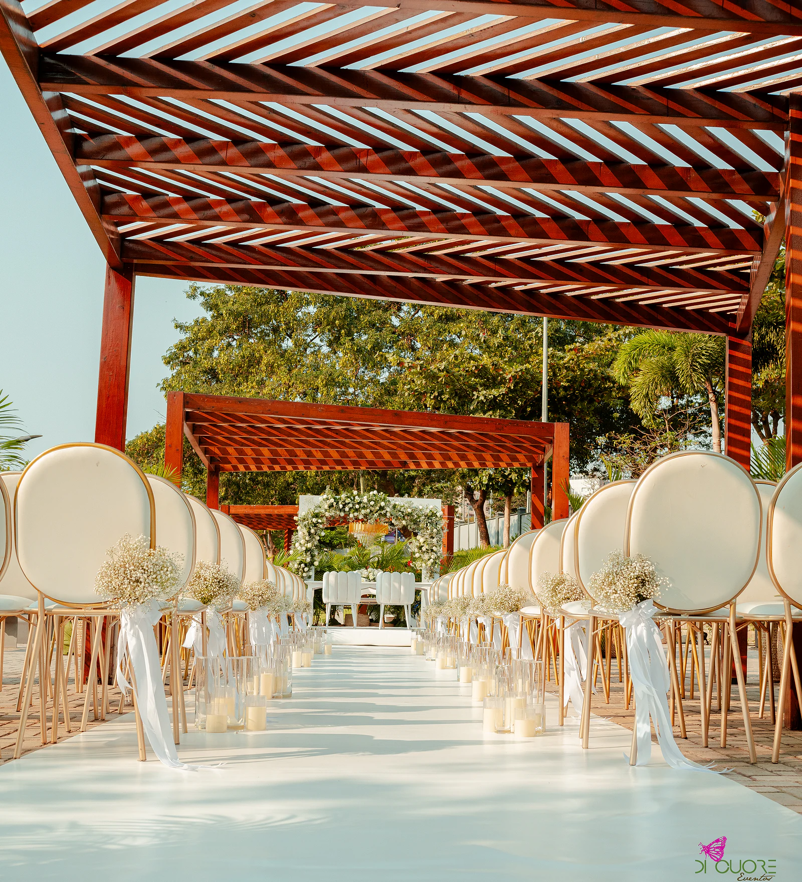 Passadeira branca sob uma pérgola de madeira, entre fileiras de cadeiras de costas redondas com ramos de gisófila e velas em cilindros de vidro