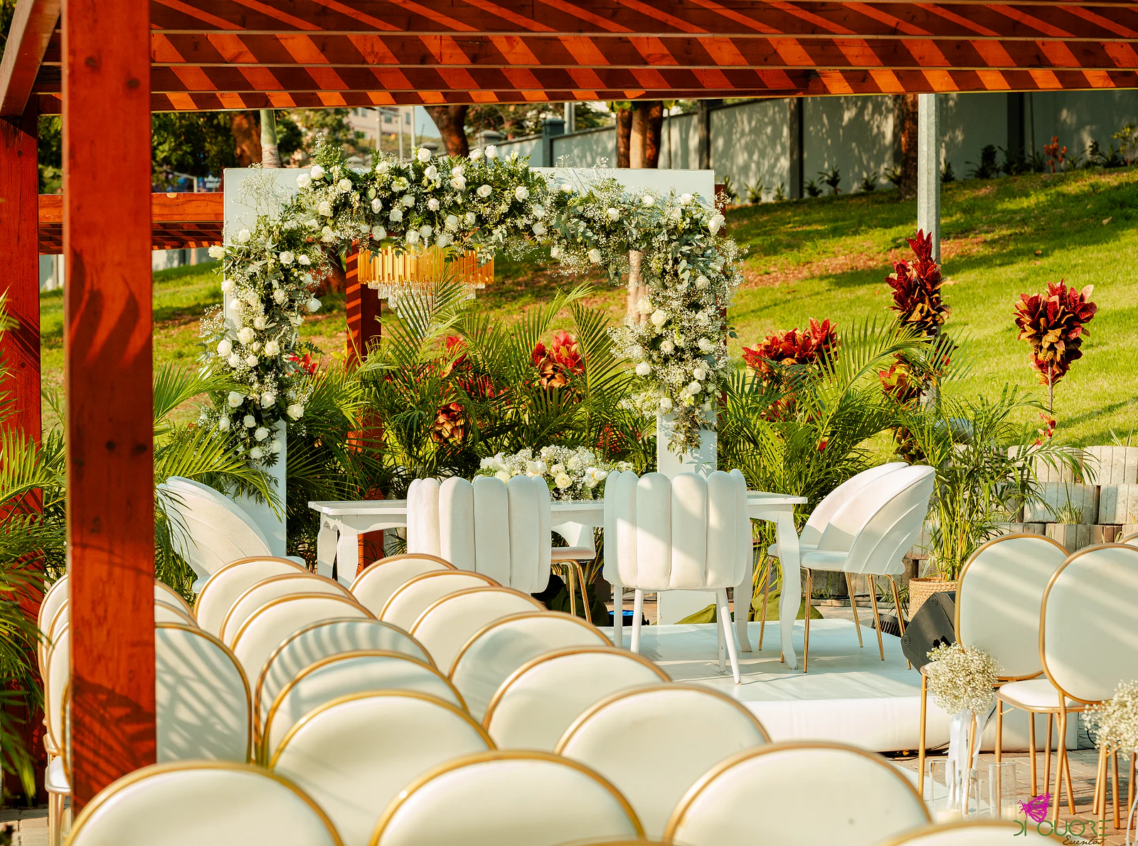 Arco de flores brancas e folhagem sobre um estrado branco com mesa e cadeiras, entre plantas de folha vermelha e uma pérgola de madeira, com cadeiras brancas de debrum dourado em primeiro plano