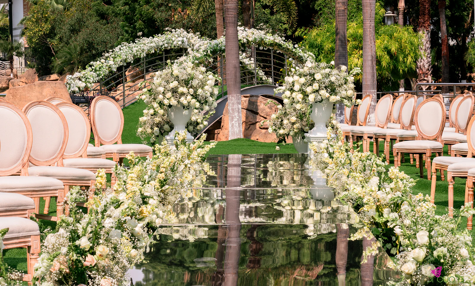 Passadeira de espelho entre cadeiras de madeira cor de pêssego e urnas de flores brancas, com uma ponte de jardim coberta por um arco de flores ao fundo
