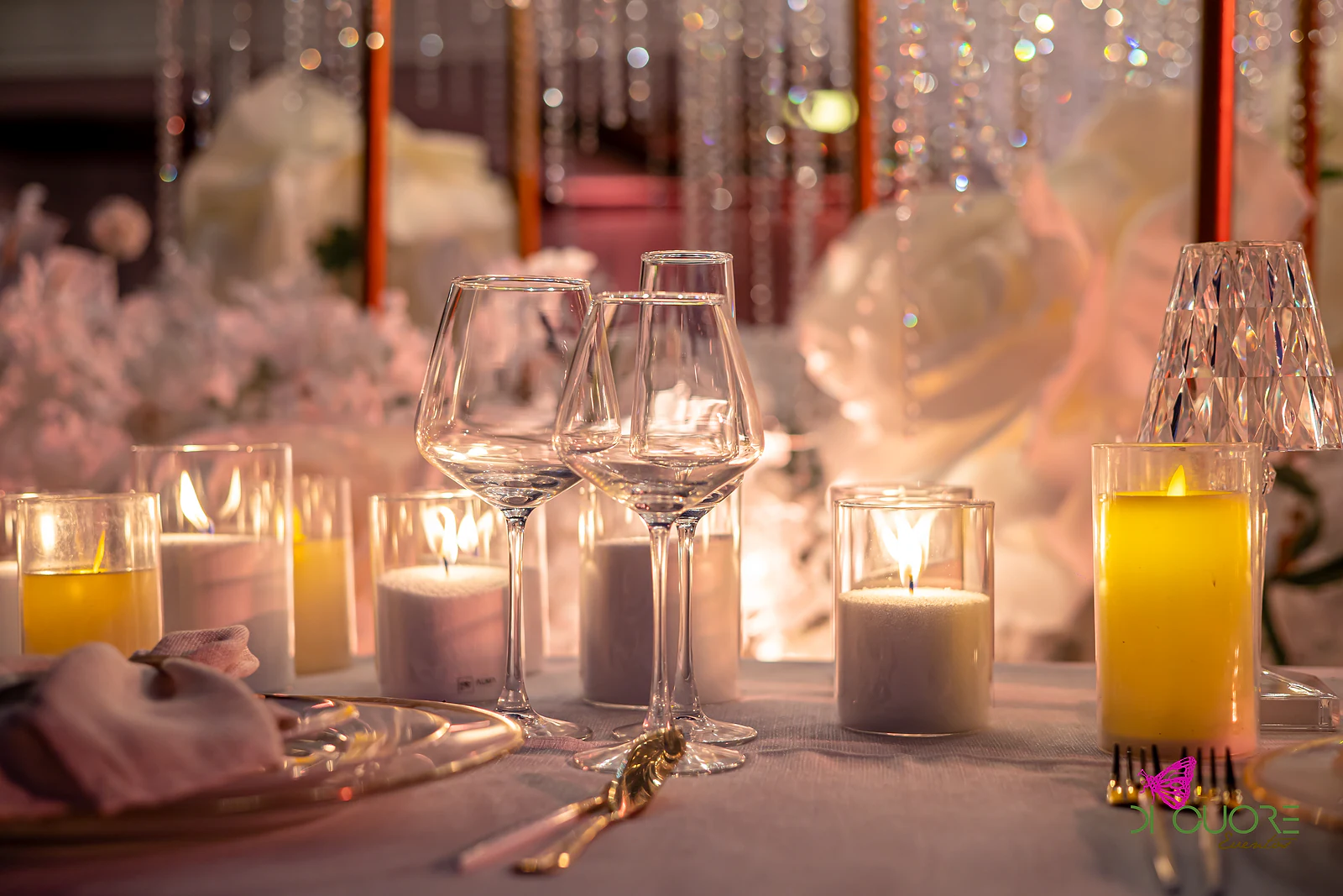 Copos de cristal, velas em copos de vidro e um candeeiro de cristal diante de fios de cristal e rosas brancas desfocadas