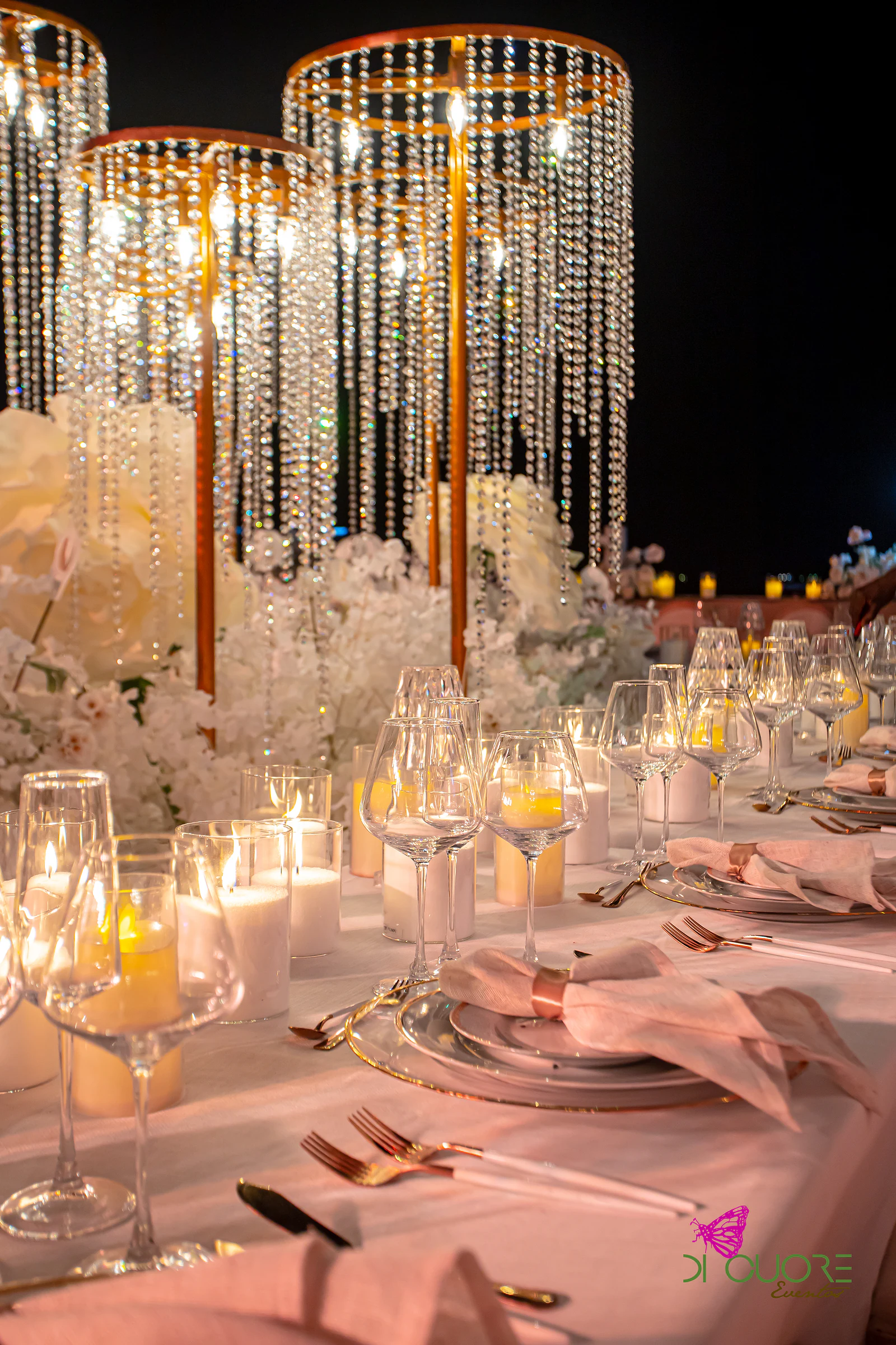 Cascatas de cristal sobre flores brancas e velas, com guardanapo cor-de-rosa com argola dourada sobre prato de rebordo dourado