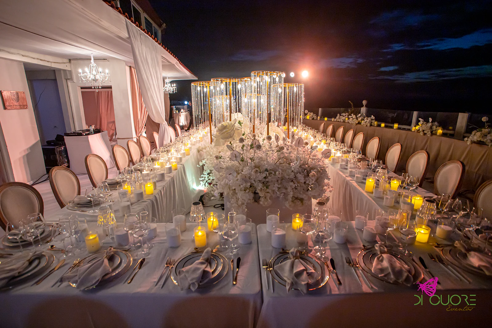 Mesa em rectângulo com um centro de flores brancas e estruturas douradas de cristal ao fundo, lugares postos, velas e cadeiras de medalhão