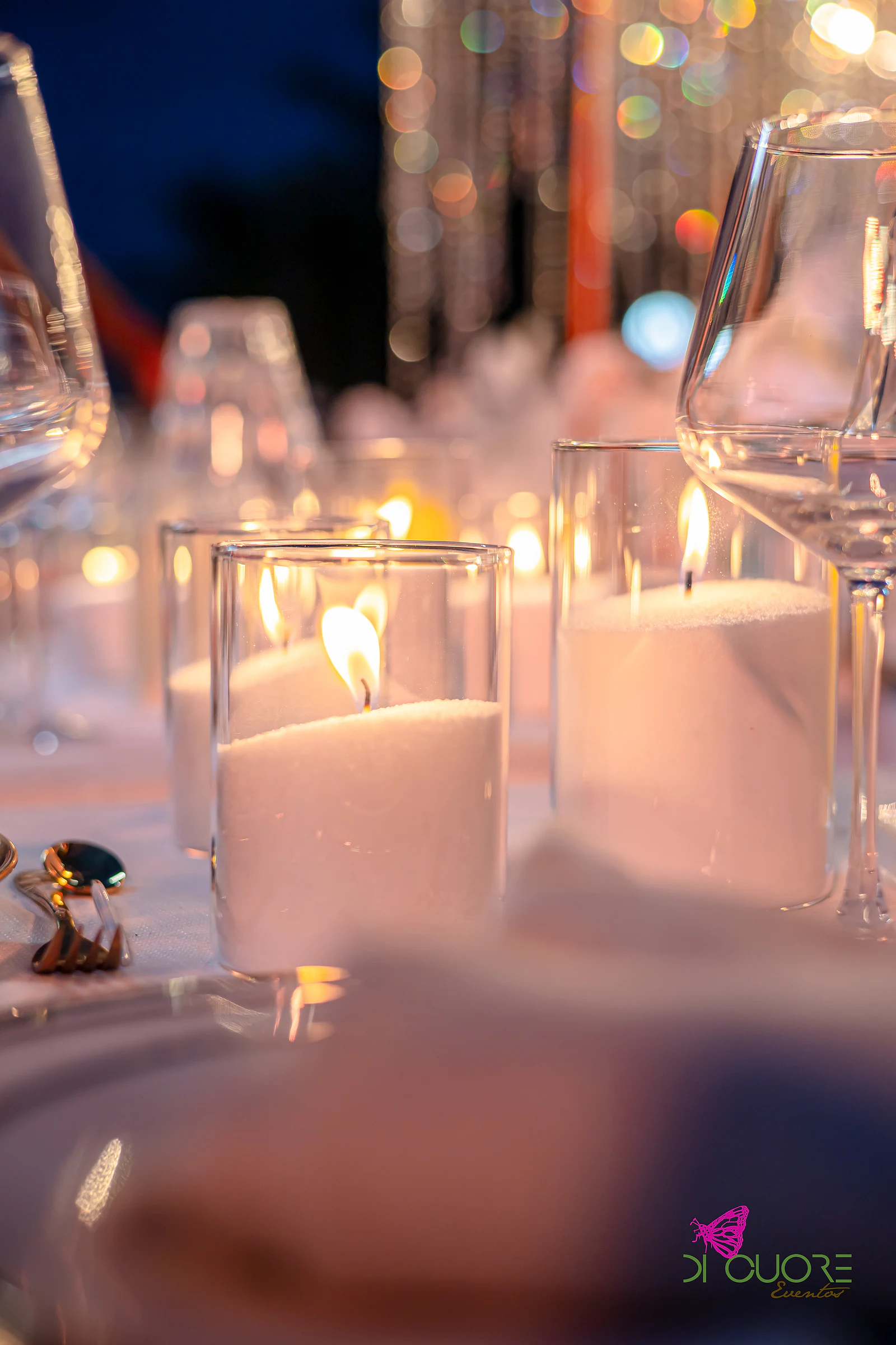 Velas em copos de vidro e copos de cristal sobre a mesa, com fios de cristal desfocados ao fundo