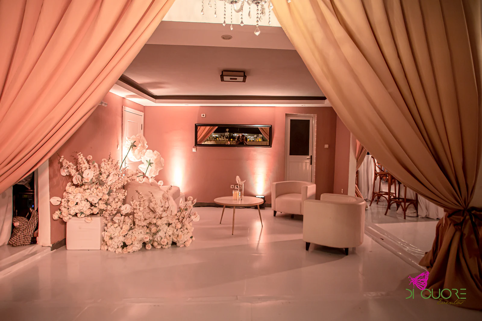 Lounge com sofá e poltronas brancas, mesa de centro dourada e massa de flores brancas, enquadrado por cortinados cor de pêssego