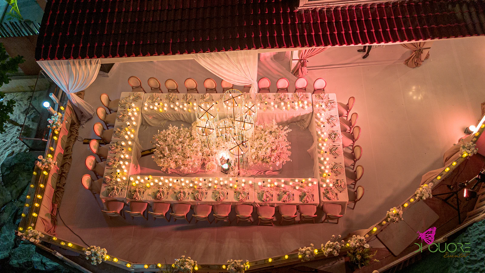 Vista de cima de uma mesa corrida em rectângulo, com velas, rosas brancas e estruturas douradas ao centro, num terraço iluminado por luzes quentes