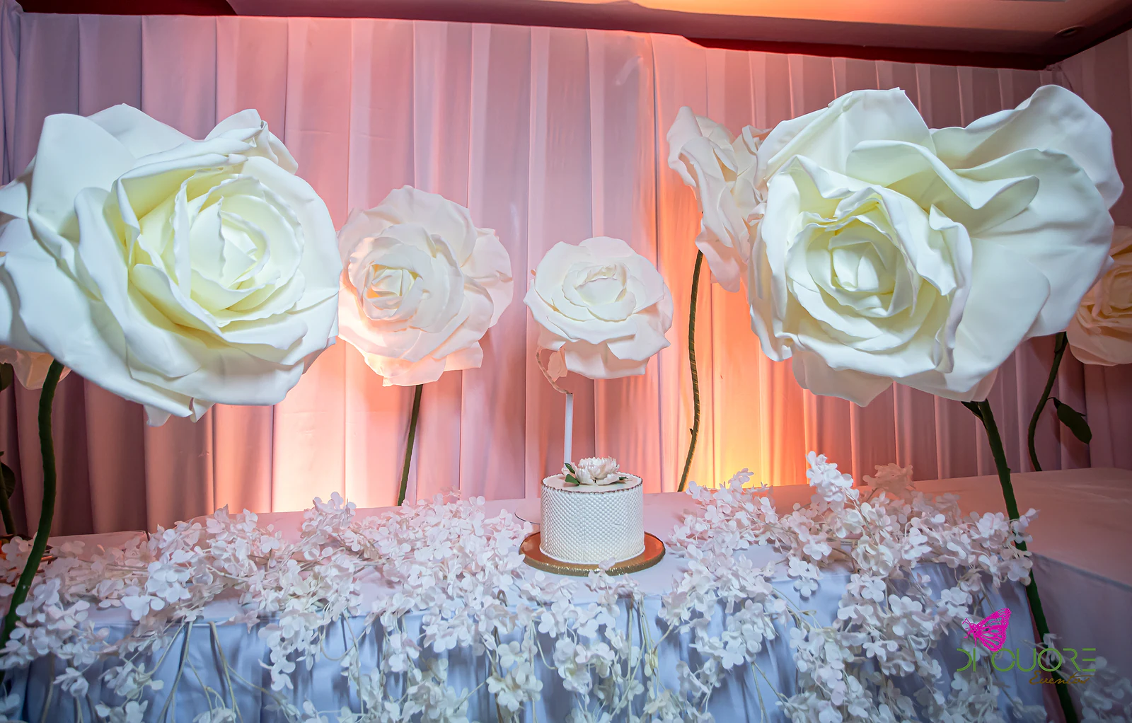 Bolo branco sobre mesa coberta de flores brancas, entre rosas gigantes e diante de cortinados rosados