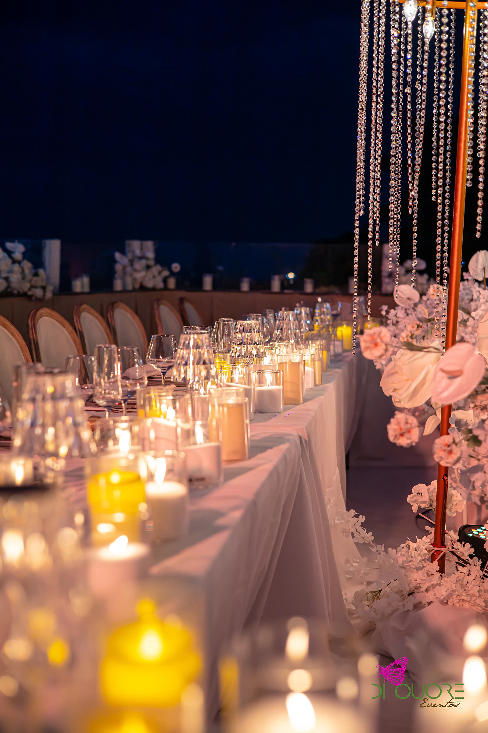 Mesa comprida iluminada por velas, em perspectiva, com cadeiras de medalhão ao fundo e uma estrutura dourada de cristal à direita