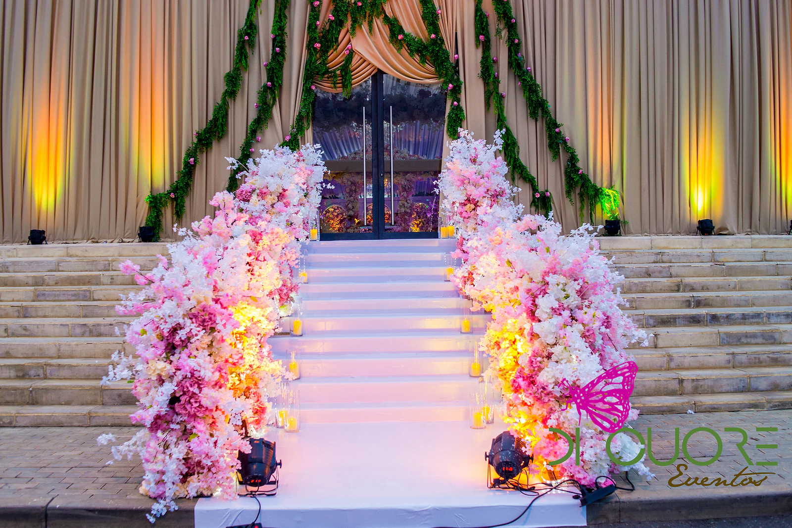 White steps lined with arrangements of pink and white flowers, with candles and spotlights, leading to the entrance of the sand-coloured marquee.