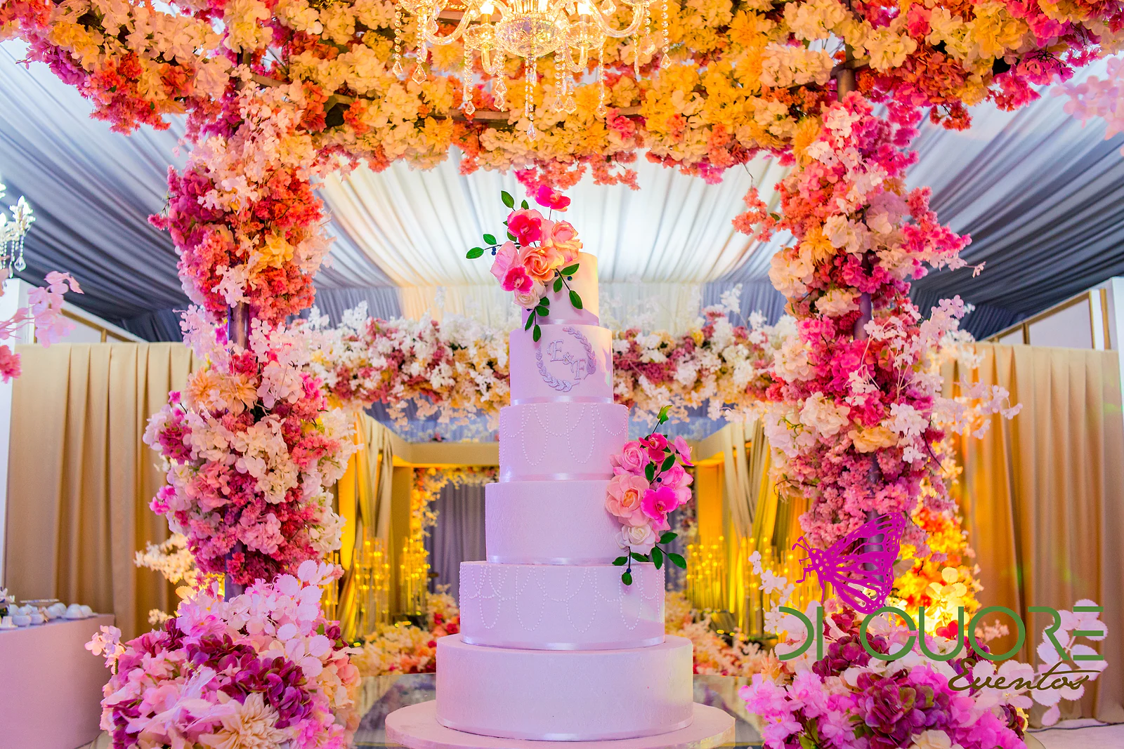 White multi-tiered cake with sprays of pink roses and monogrammed initials, beneath a canopy of pink and orange flowers with a chandelier.