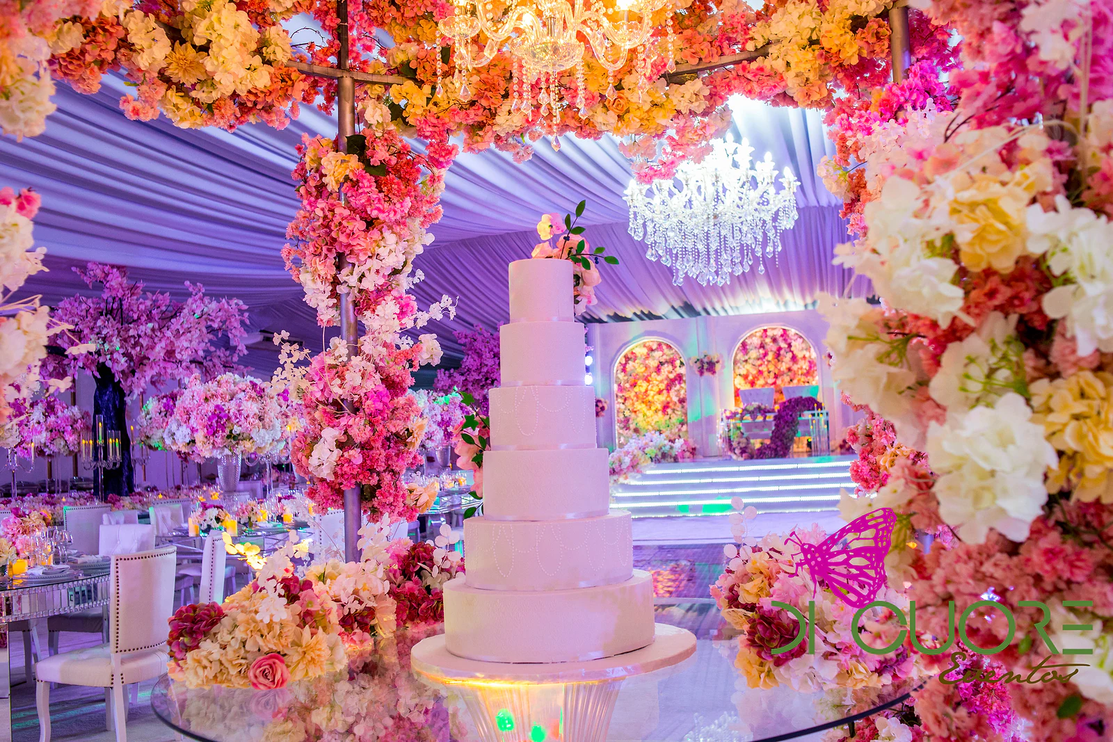 Multi-tiered cake beneath the floral canopy and chandelier, with the lit stage at the far end of the hall.