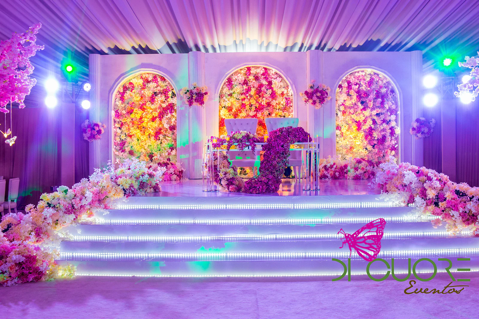 Stage with three arched niches covered in flowers, lit steps and the main table with a cascade of purple blooms, beneath the draped ceiling.