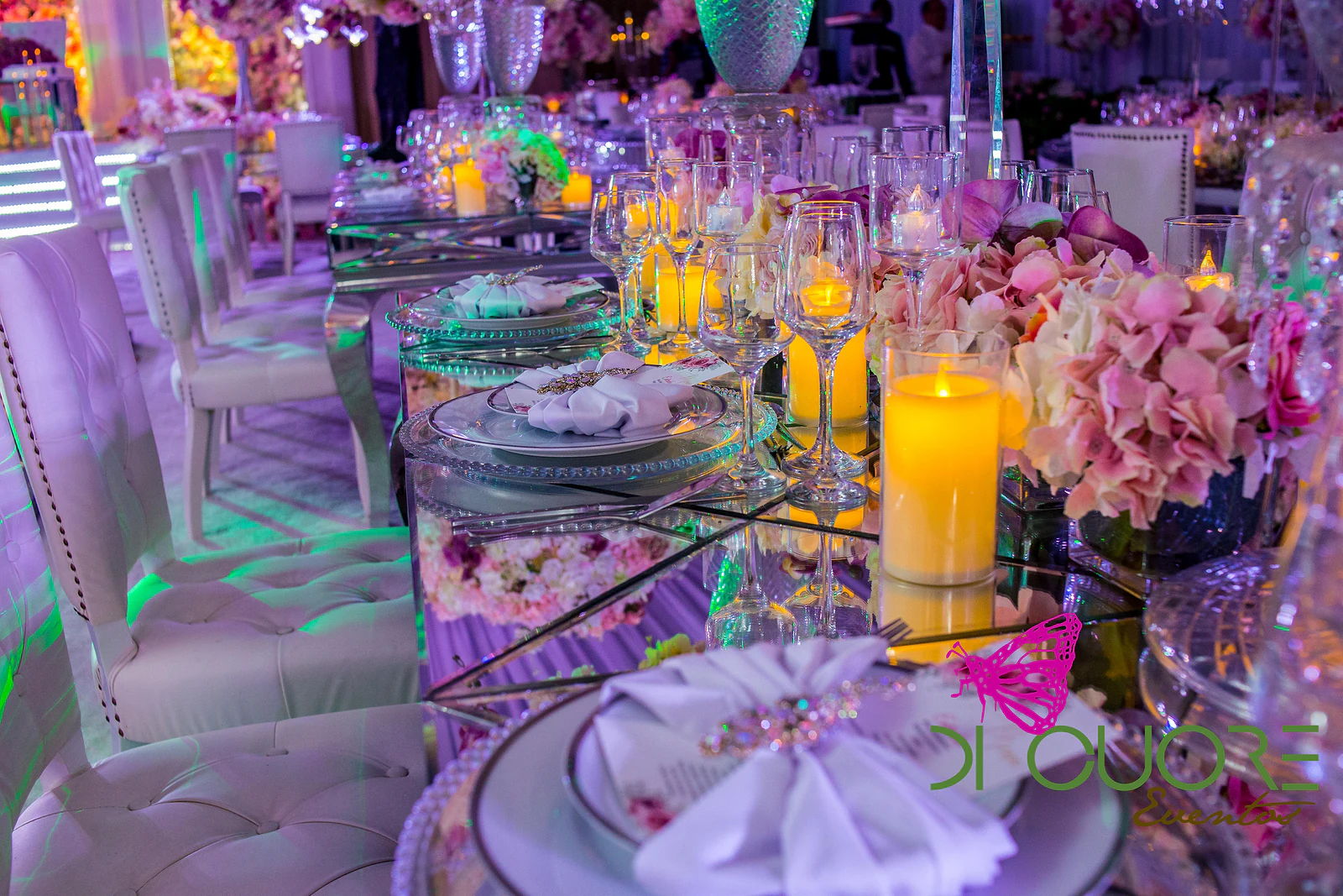 Mirrored table with candles, crystal glasses, plates with beaded chargers and pink flower arrangements, beside white tufted chairs.