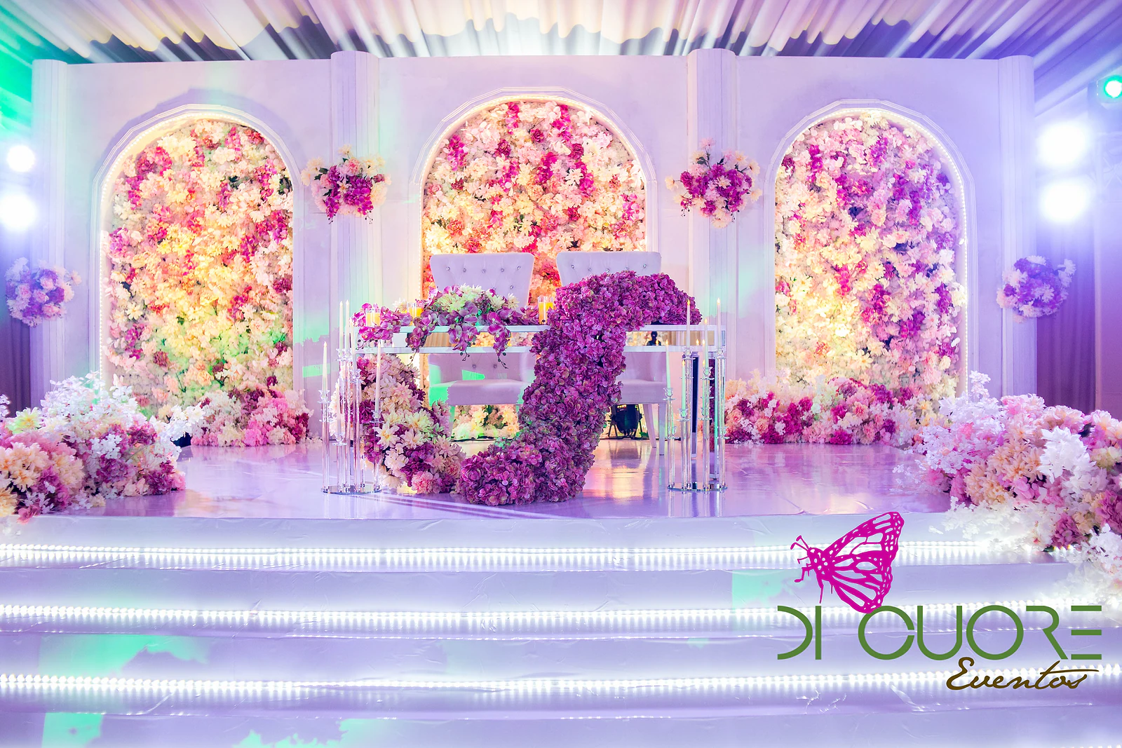 Mesa dos noivos com cadeiras estofadas e uma cascata de flores roxas em primeiro plano, diante de arcos de flores cor-de-rosa e amarelas, sob um tecto de cortinados brancos