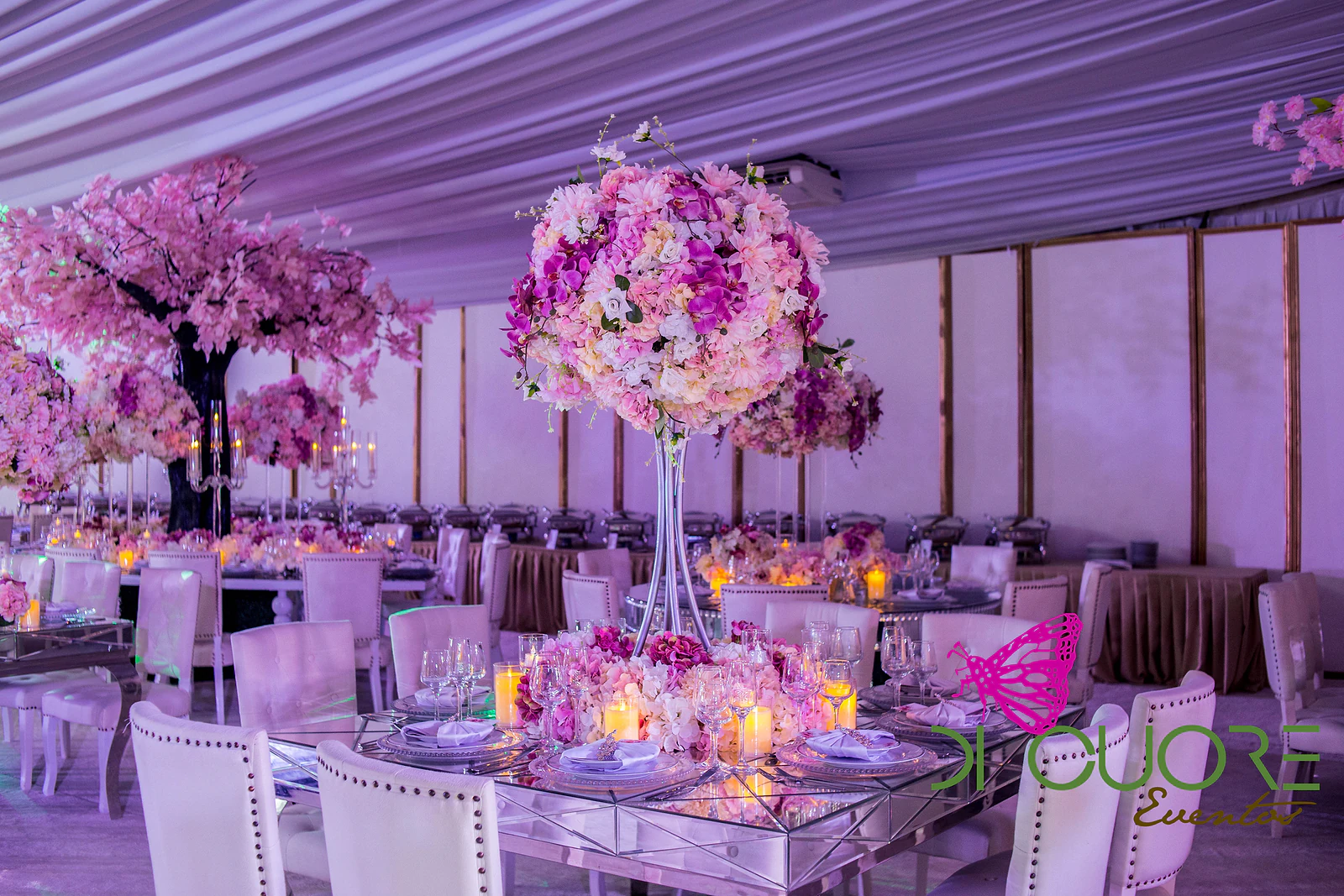 Tall centrepiece of pink and purple flowers on a mirrored table, with a pink blossom tree behind.