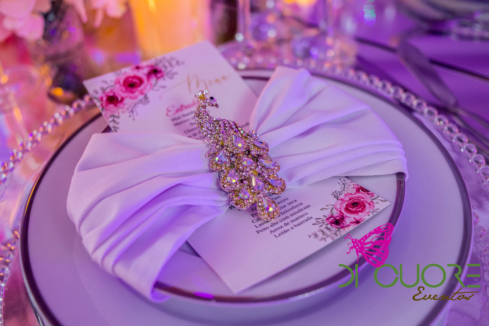 Plate with a white napkin held by a crystal peacock brooch and a menu card decorated with pink flowers.