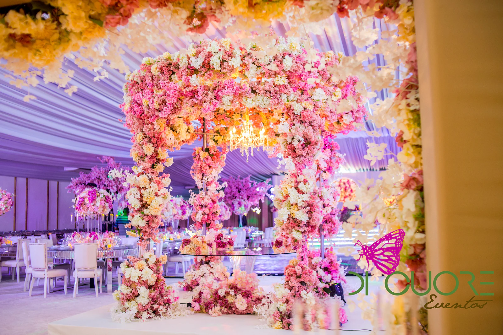 View through pink flowers to a flowered gateway and the hall beyond, between golden drapes and crystal chandeliers.