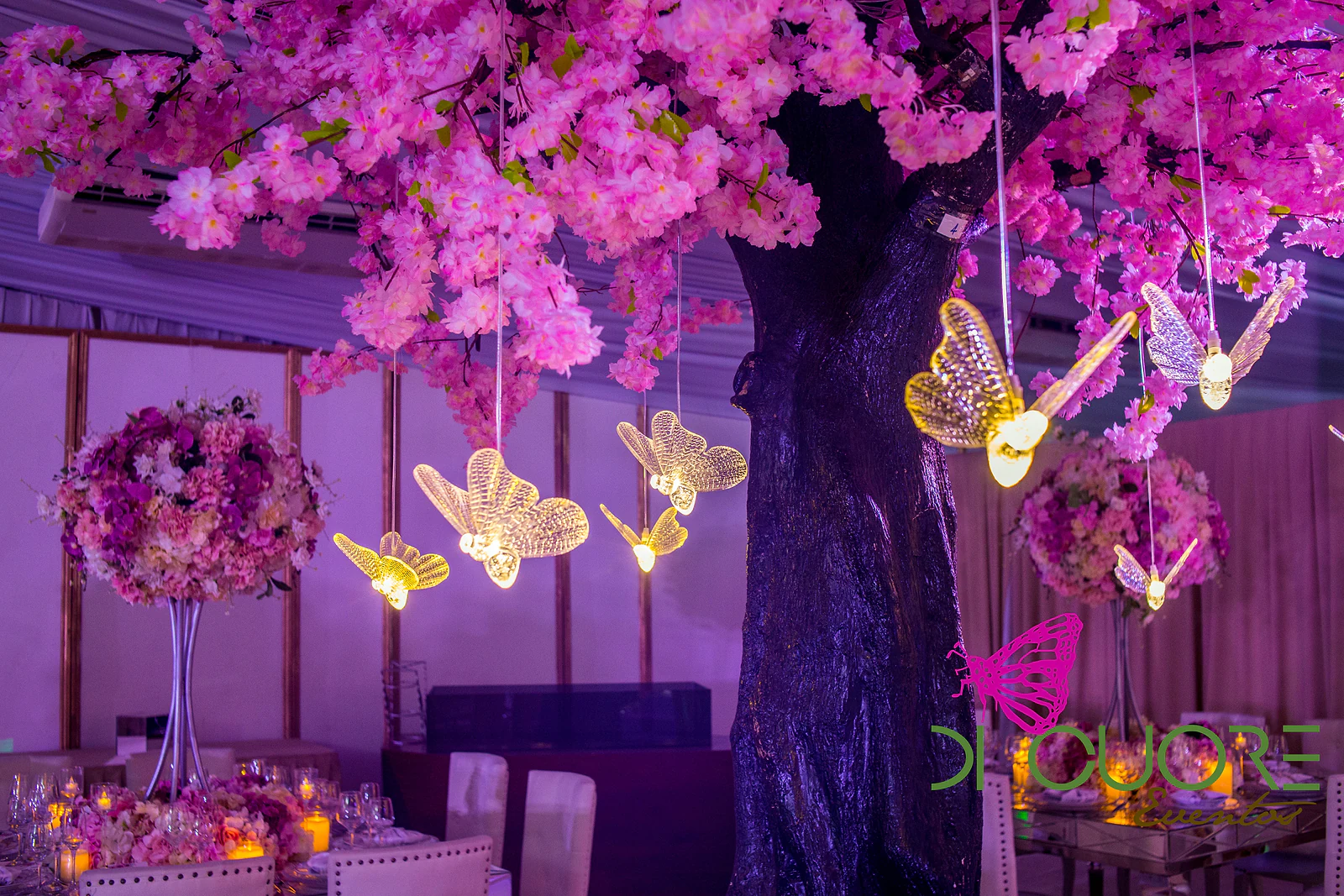 Pink blossom tree hung with lit golden butterflies, above tables with flower centrepieces.