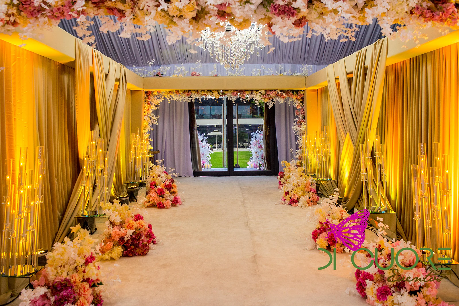 Corridor of crossed golden drapes, with crystal candelabra and sprays of flowers on the floor, opening onto a glass door.