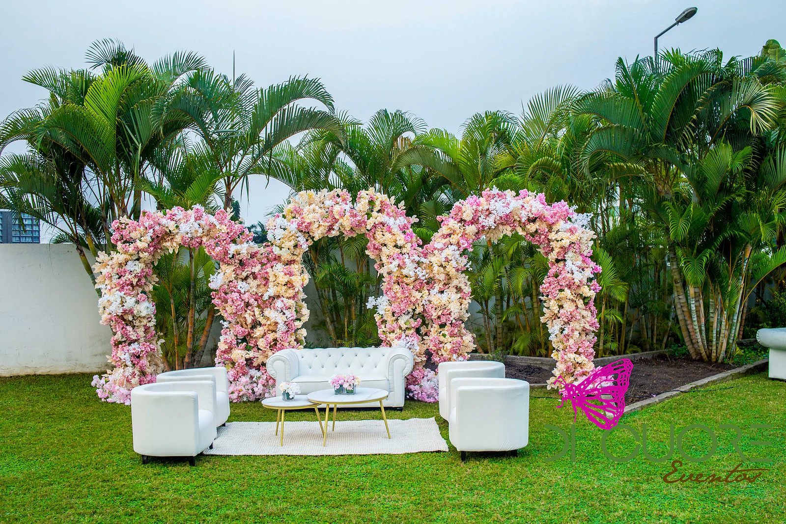 Three arches of pink and peach flowers on a lawn, with white sofas and a side table, beside palm trees.