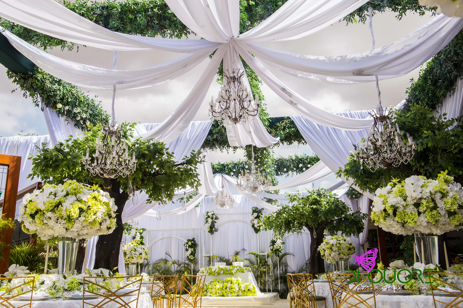 Ceiling of white drapes opening out like a star, with crystal chandeliers, foliage and trees above hydrangea arrangements