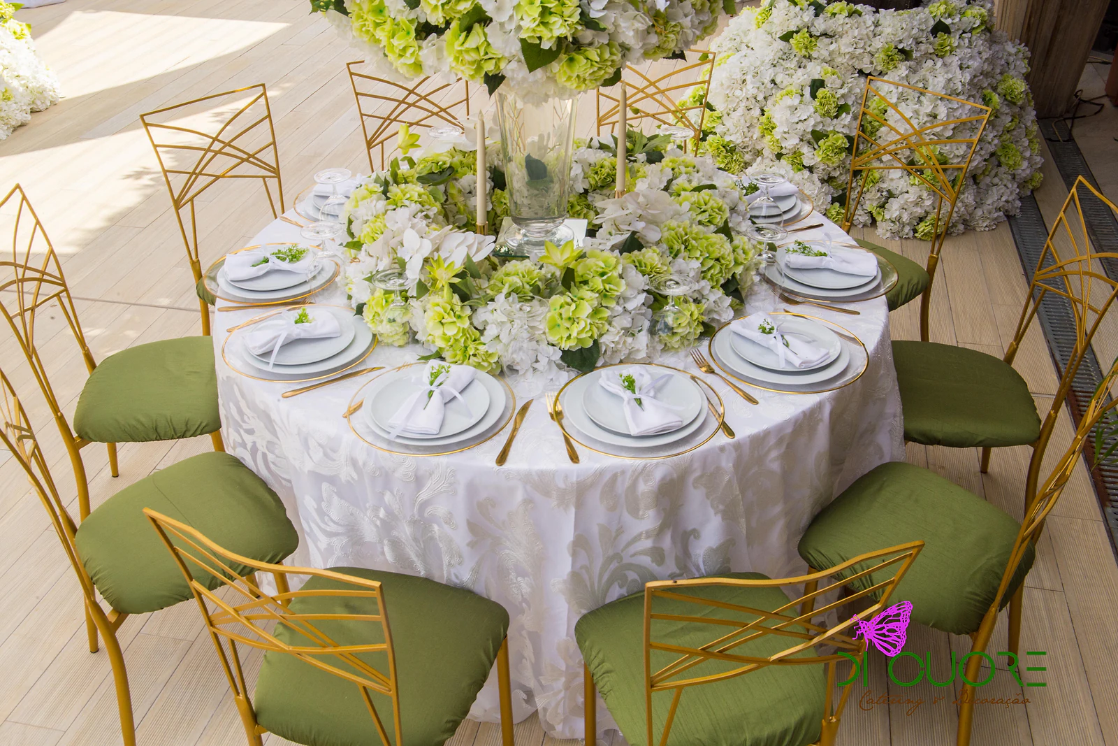 Round table in a white damask cloth, with a centrepiece of hydrangeas and candles, gold-rimmed plates and gold chairs with green seats