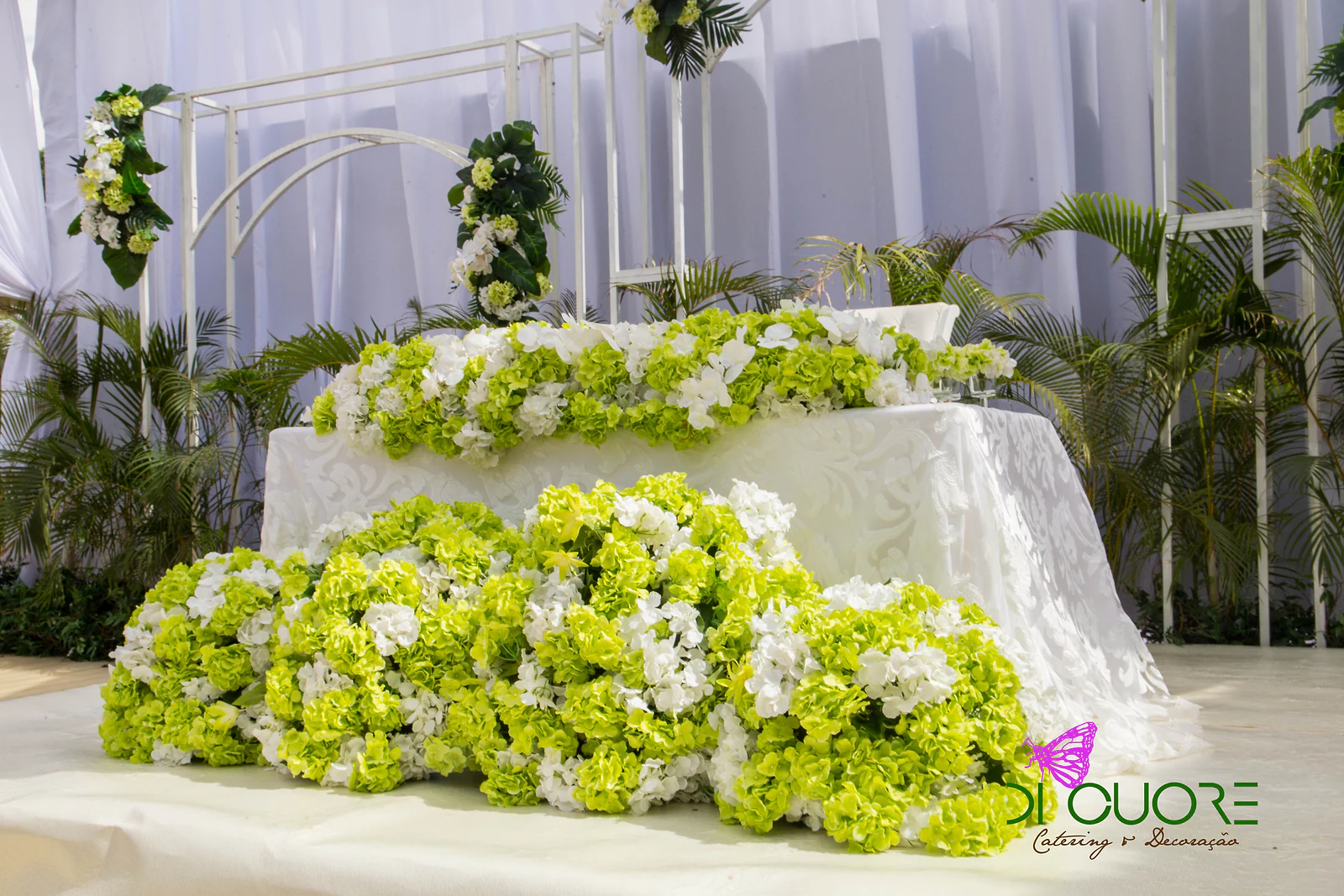 Main table in a white cloth with a band of lime-green and white hydrangeas and large masses of flowers in front, before white arches and palms