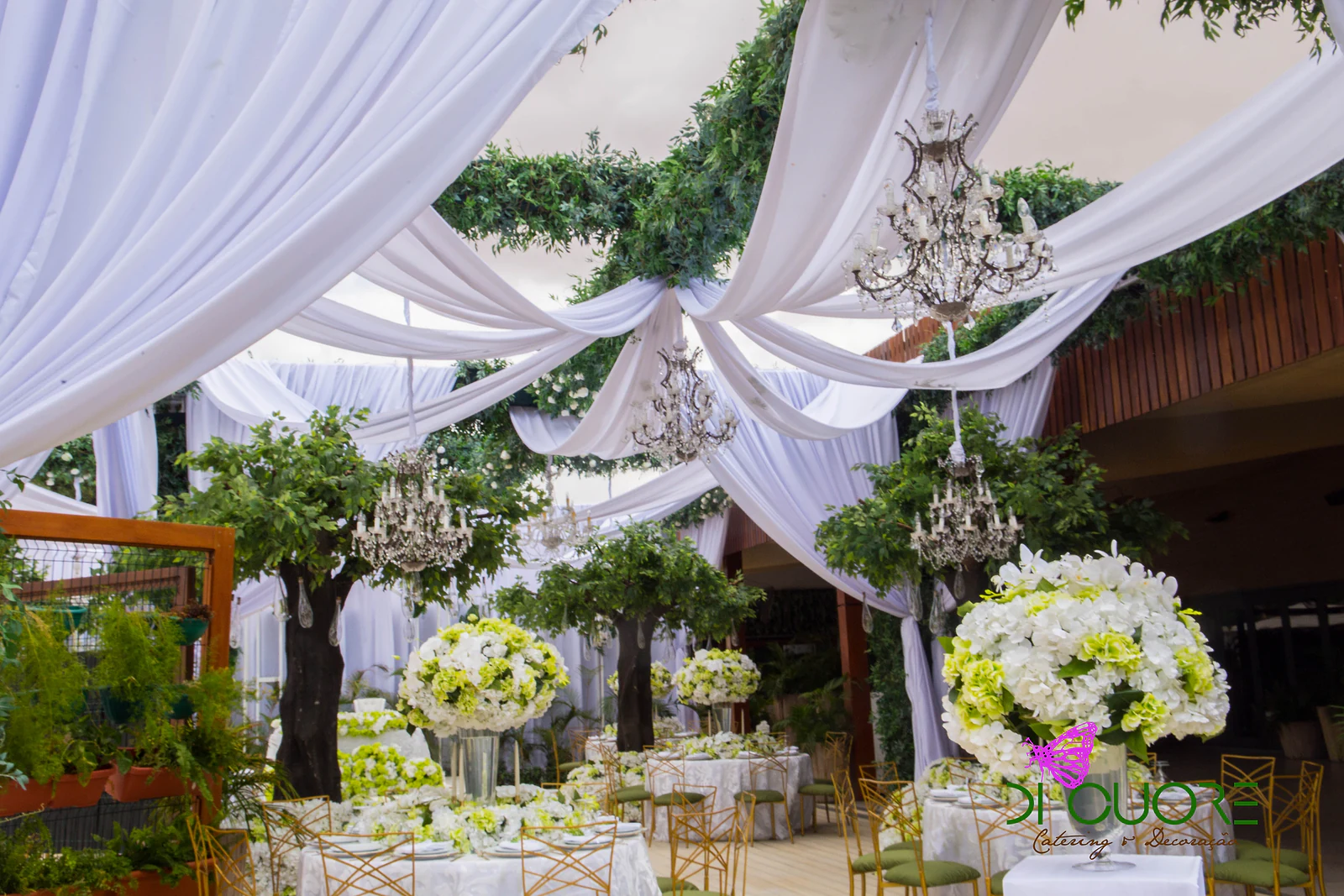Wide view of the space, with chandeliers above the trees, tall hydrangea arrangements and laid tables beneath a ceiling of drapes