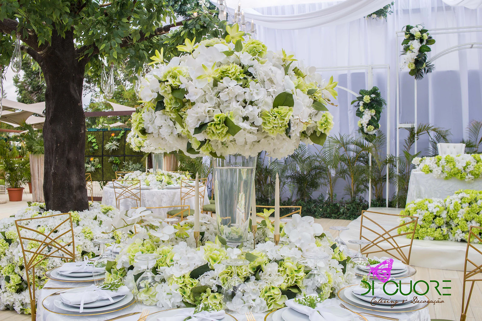 Tall arrangement of white and lime-green hydrangeas in a glass vase with candles on the laid table, with a tree behind