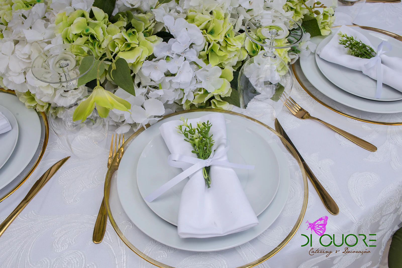 Gold-rimmed plate with a ribbon-tied napkin and a sprig of green, among gold cutlery and hydrangeas