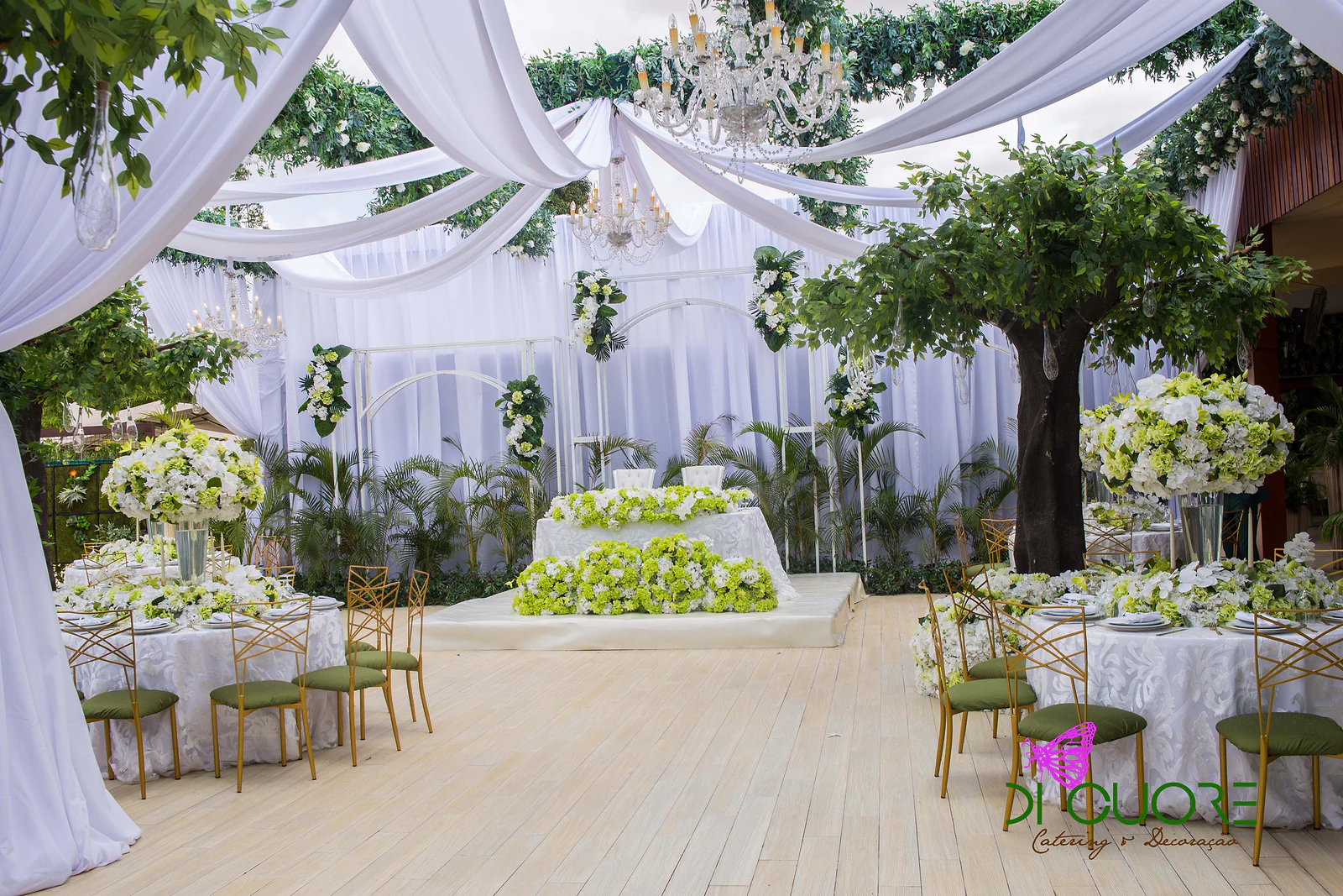 Pale wooden floor between round tables, with the main table at the far end, white drapes, chandeliers and trees