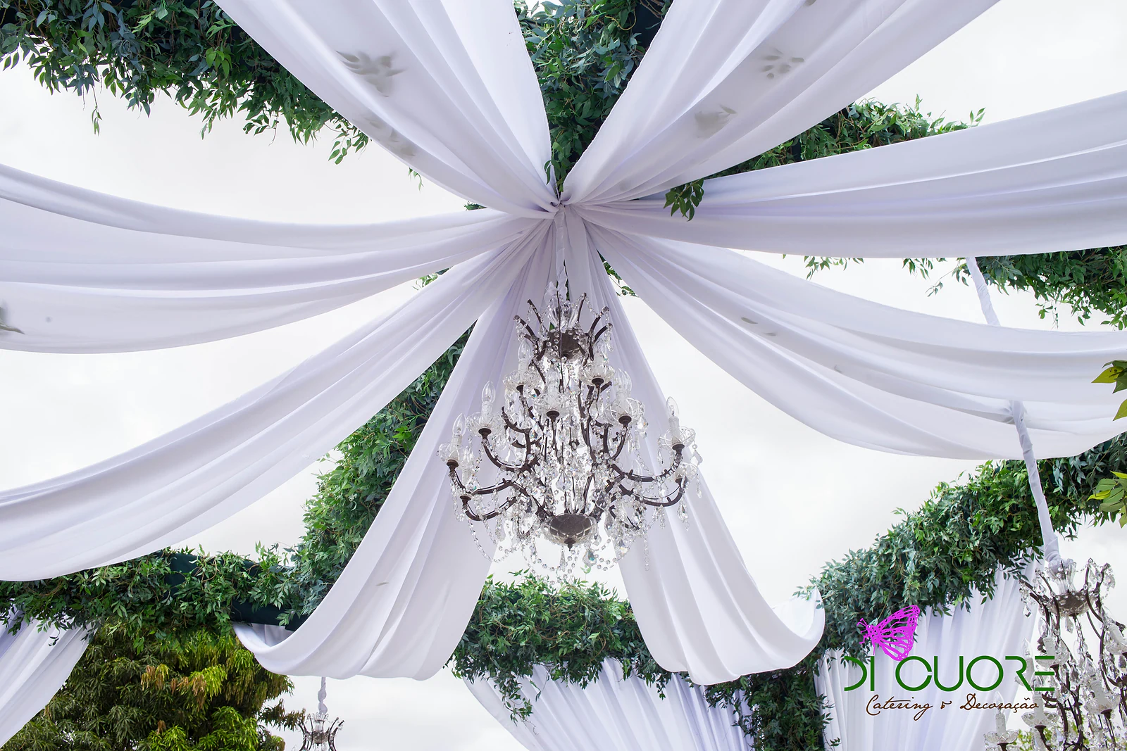 Crystal chandelier hanging at the centre of a ceiling of white drapes, among foliage