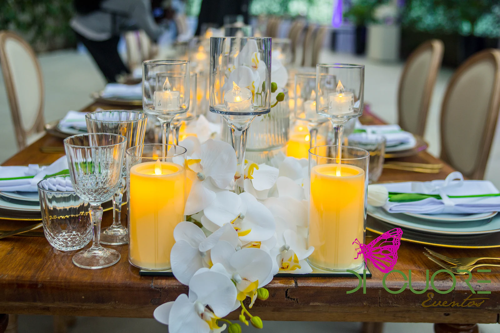 Velas em copos de vidro, taças de cristal e orquídeas brancas sobre um espelho, junto a um prato com rebordo dourado.