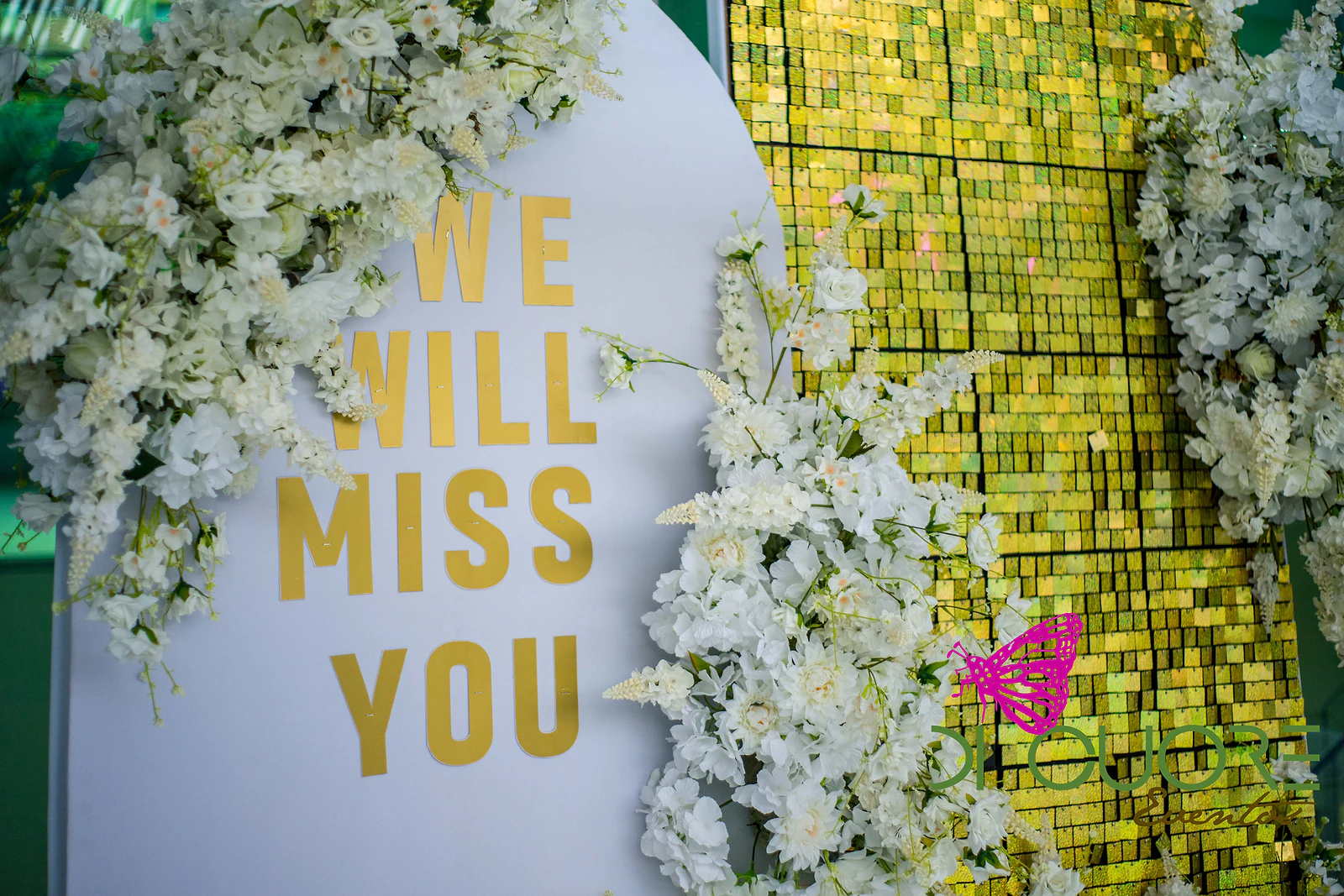 Painel branco com a mensagem “WE WILL MISS YOU” em letras douradas, rodeado de flores brancas, junto a uma parede de lantejoulas douradas.
