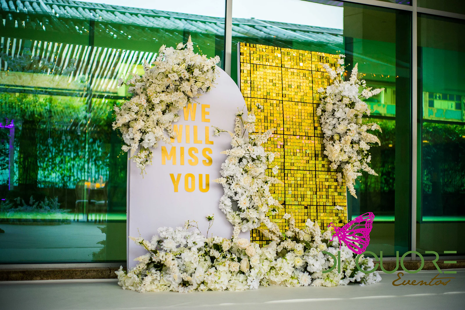 Painel em arco com a mensagem “We will miss you”, flores brancas e uma parede de lantejoulas douradas diante de uma fachada de vidro.