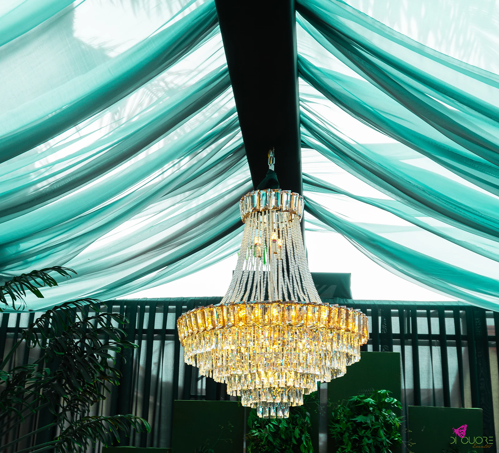 Lustre de cristal com pendentes dourados sob o tecto de seda verde-água, com folhagem em vasos e painéis verdes ao fundo
