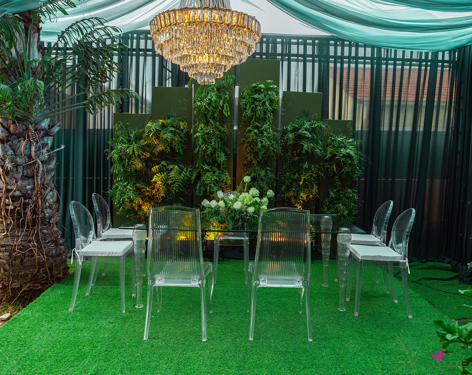 Mesa da cerimónia com cadeiras transparentes diante de um painel de folhagem e de um lustre dourado, sobre tapete verde