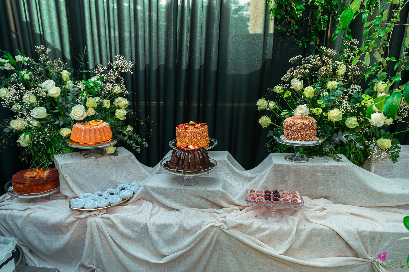 Mesa de doces com bolos e pastéis sobre toalhas de linho cru, entre ramos altos de rosas brancas e folhagem