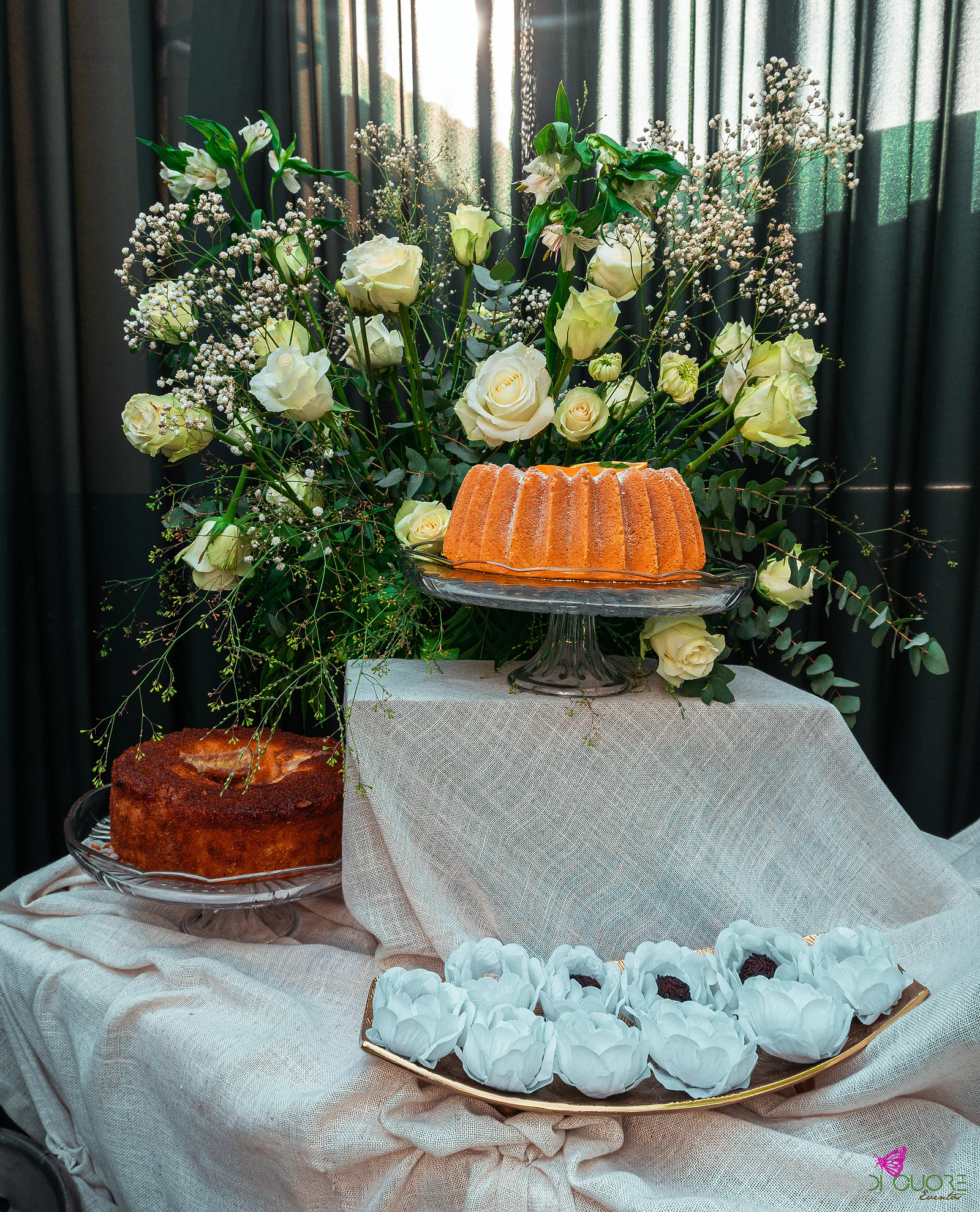 Mesa de doces sobre linho cru, com dois bolos em pé de vidro e doces brancos em forma de flor, diante de um grande arranjo de rosas claras e gipsófila