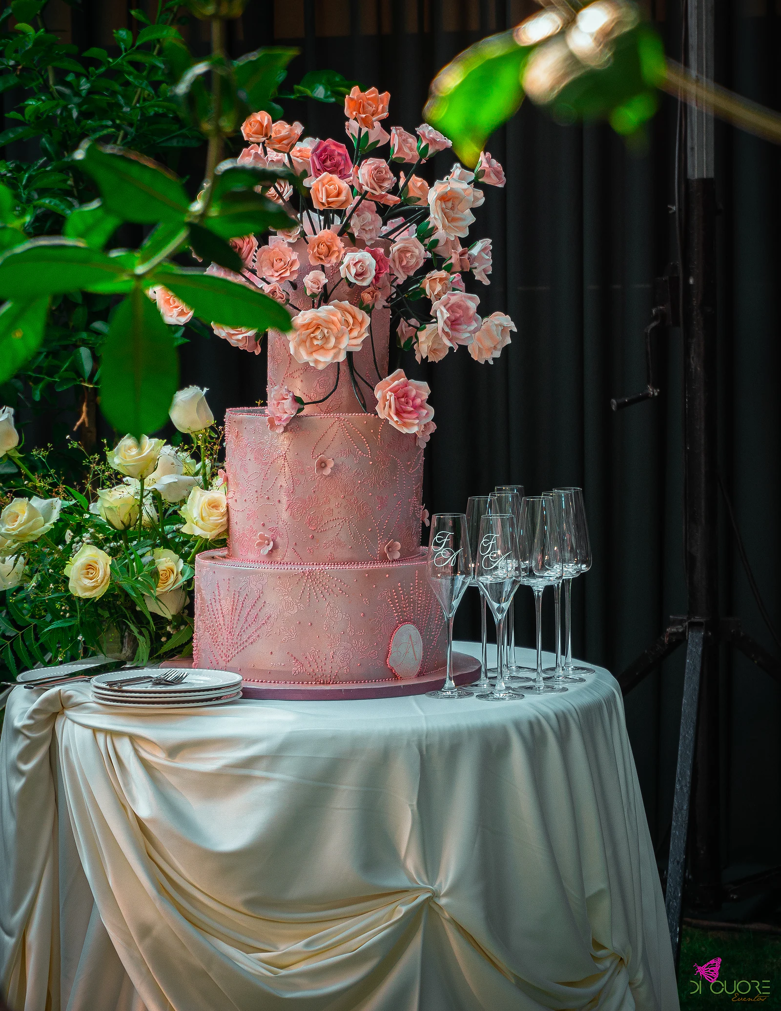 Bolo de três andares cor-de-rosa com flores de açúcar no topo, sobre uma mesa redonda de cetim marfim, junto a flores brancas