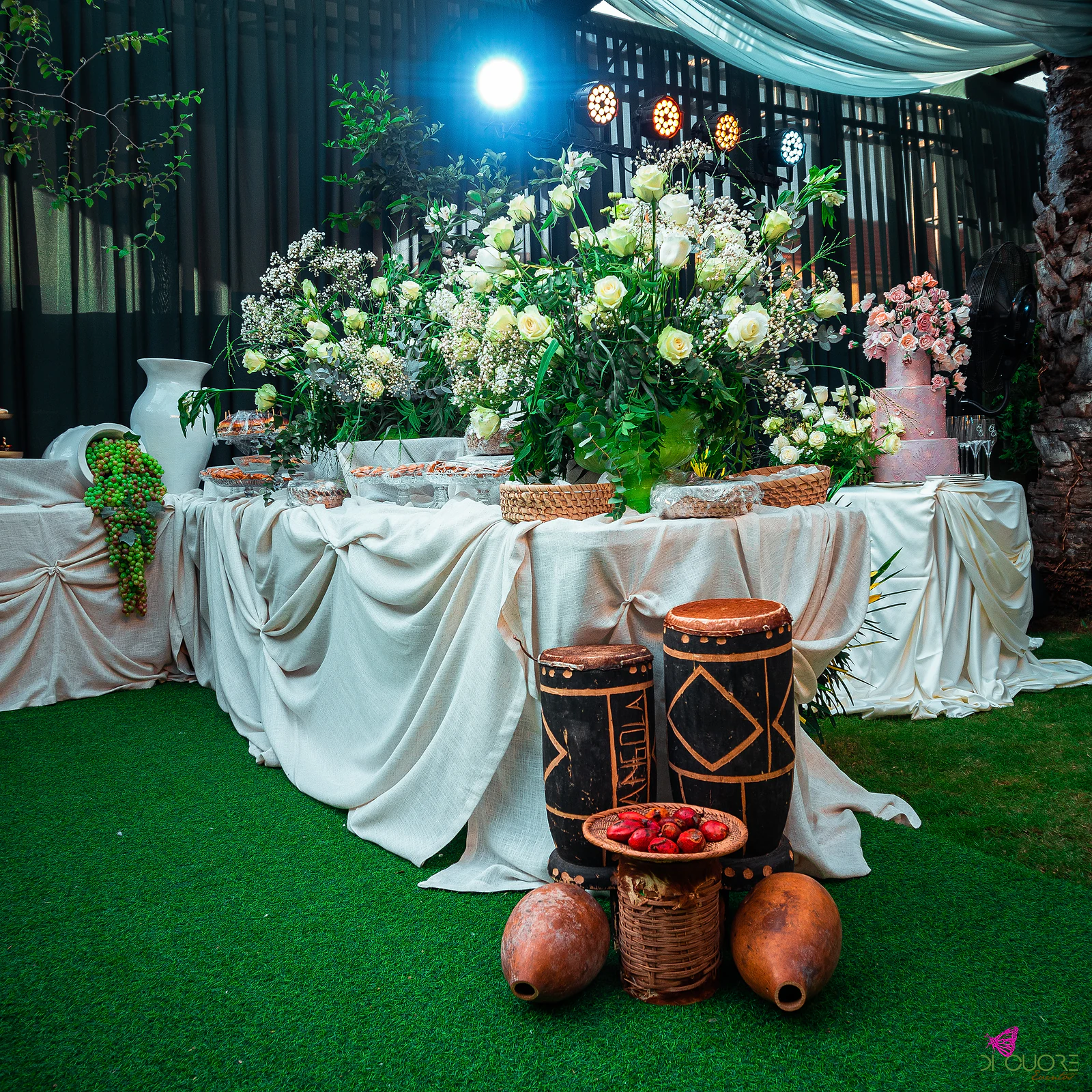 Mesa de doces sobre linho cru, com grandes arranjos de rosas claras, dois tambores decorados, cabaças e um cesto de frutos vermelhos sobre a relva, e o bolo cor-de-rosa ao fundo