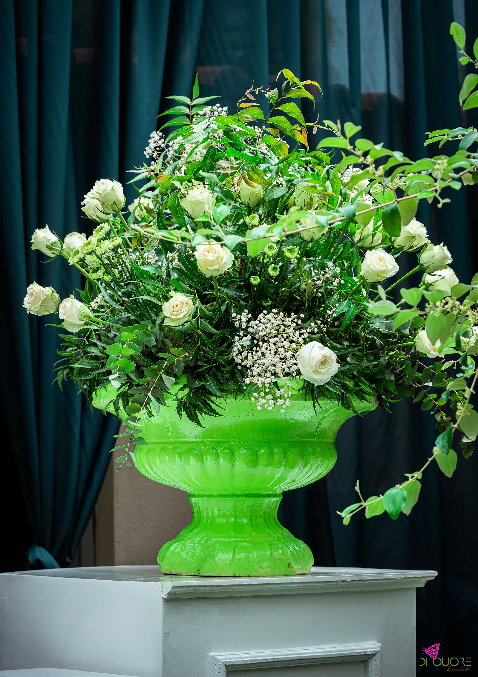 Urna verde-lima sobre um pedestal branco, com um grande arranjo de rosas brancas, gipsófila e folhagem, diante de um cortinado verde-escuro