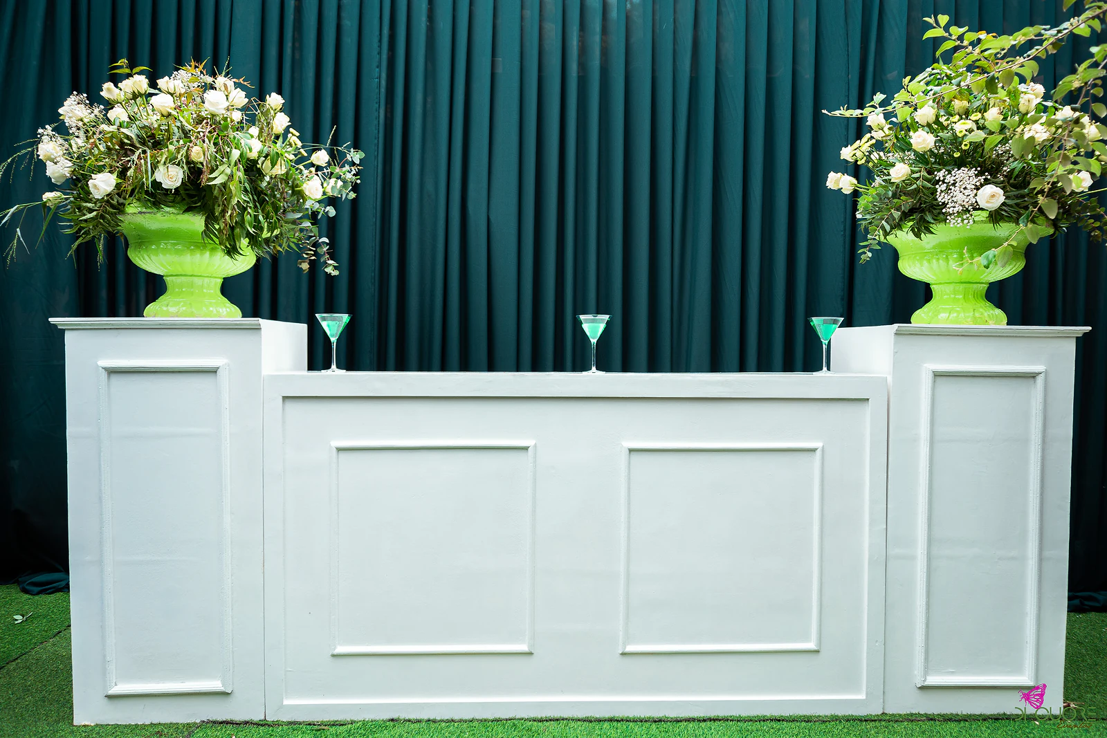 Balcão de bar branco com painéis e duas urnas verde-lima com arranjos de rosas brancas e folhagem, diante de cortinas verde-escuras