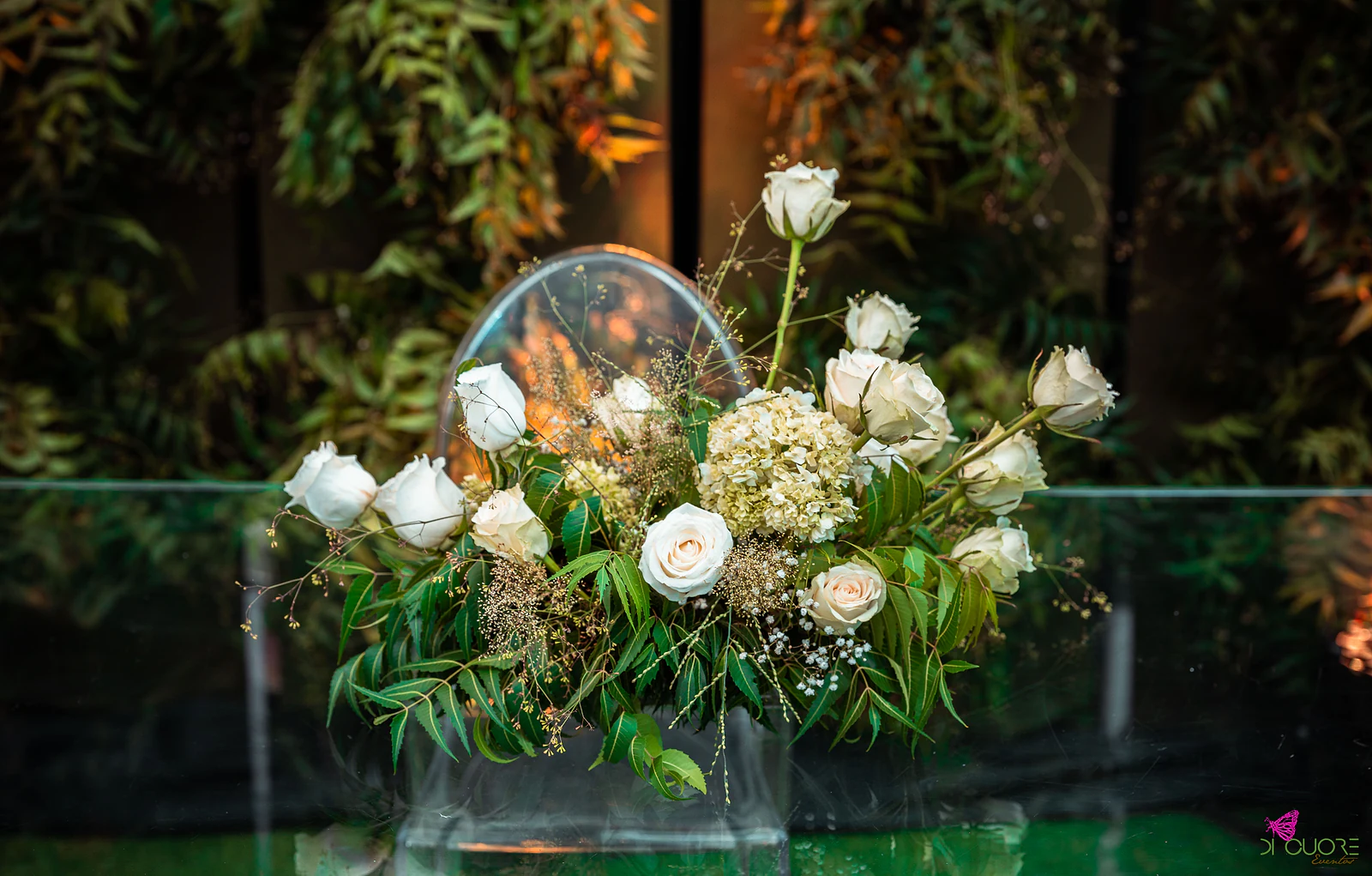 Arranjo de rosas brancas, hortênsia e folhagem sobre uma mesa de vidro, com uma cadeira transparente e folhagem iluminada ao fundo