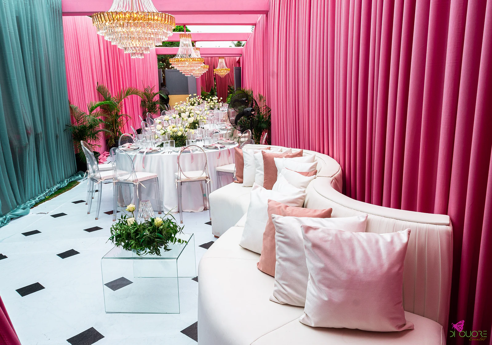 Sofá curvo branco com almofadas rosa e, ao fundo, uma mesa redonda sob lustres, entre cortinados cor-de-rosa e verde-água
