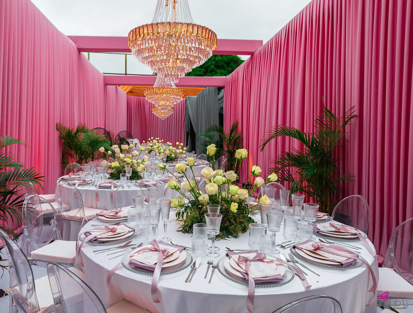 Mesa redonda posta com toalha branca, guardanapos cor de malva e um arranjo de rosas claras, sob um lustre de cristal entre cortinados cor-de-rosa