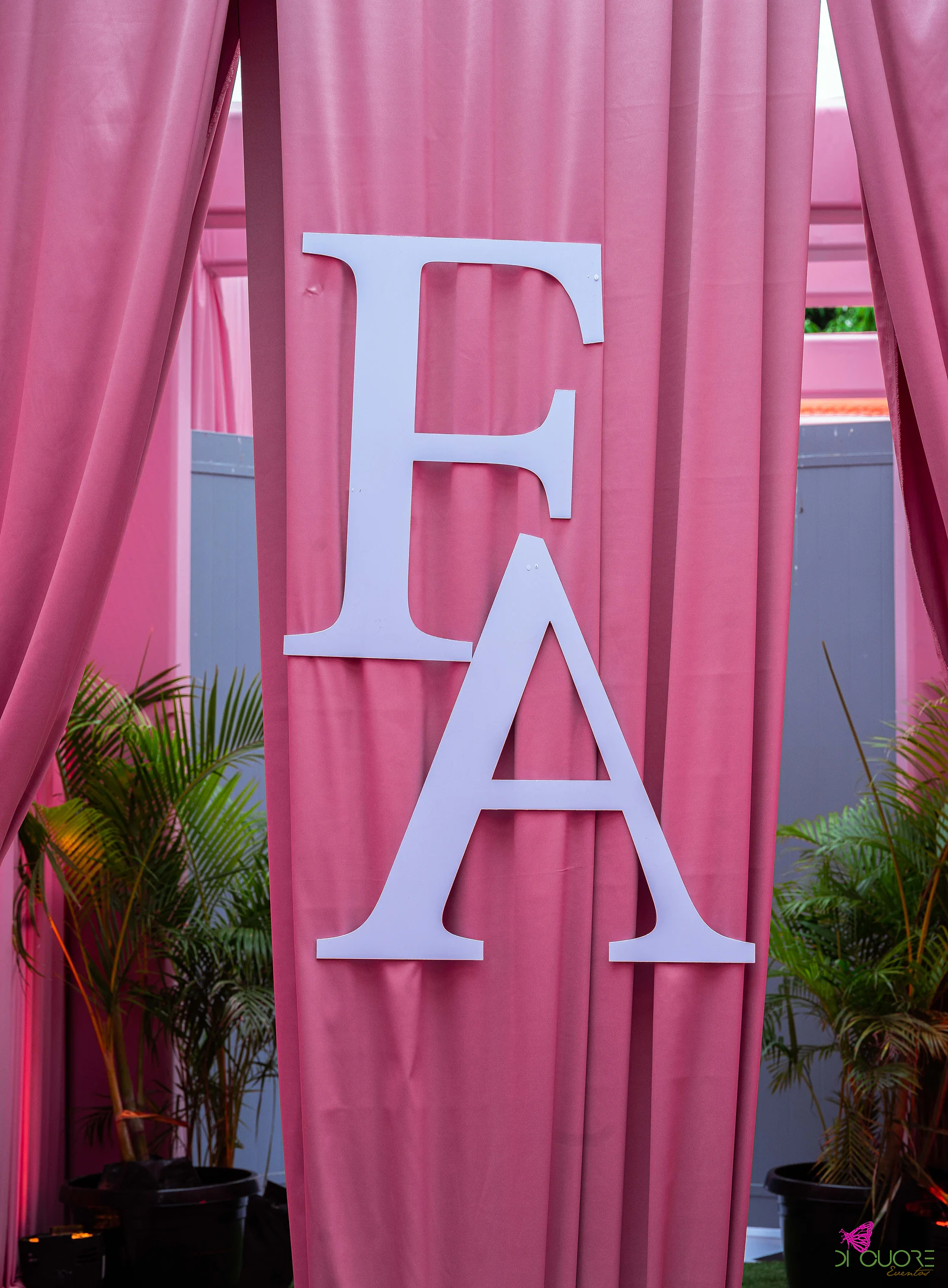 Letras F e A em branco sobre um cortinado cor-de-rosa, entre palmeiras em vasos