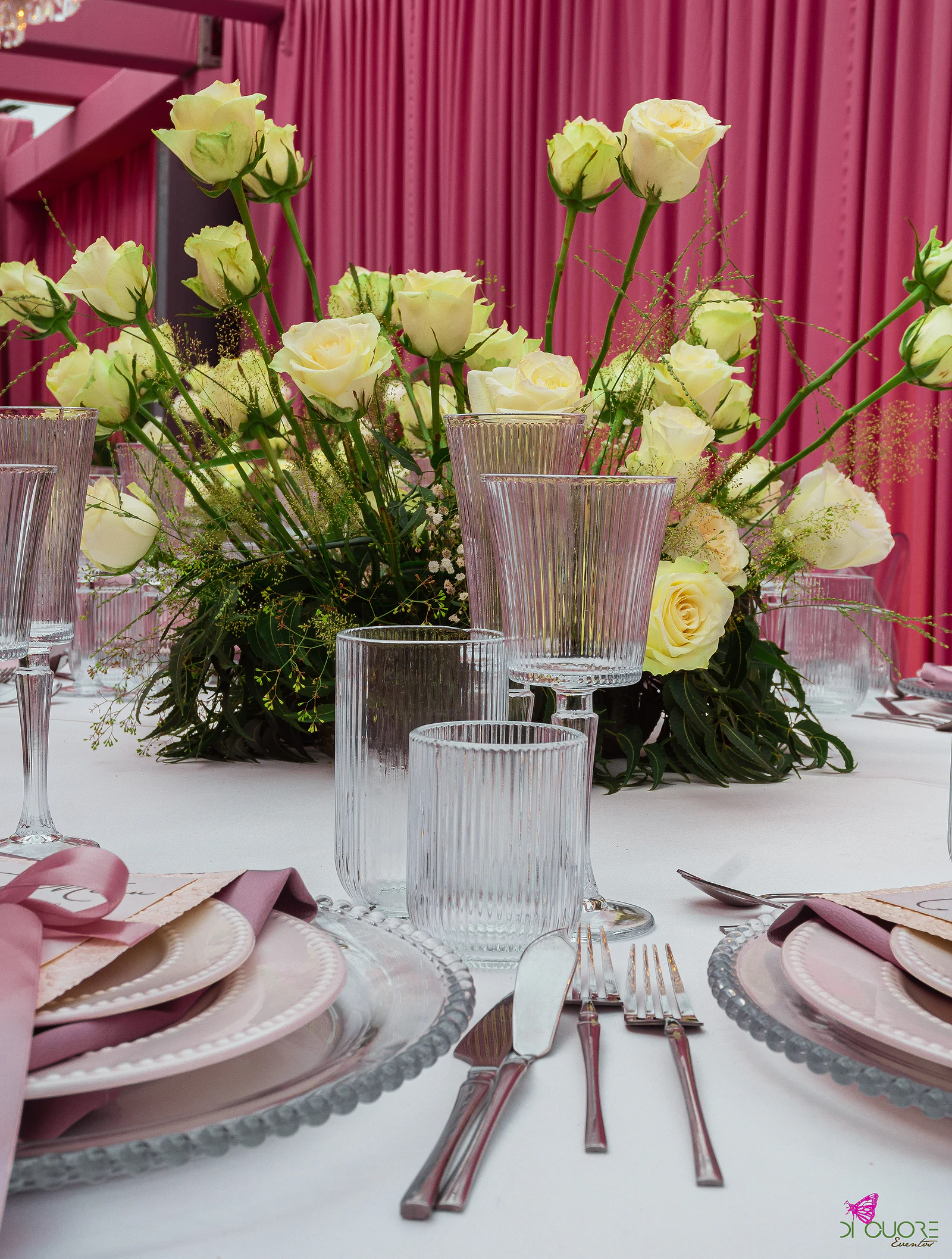 Pormenor da mesa posta com pratos de rebordo de contas, guardanapos cor de malva, copos canelados e rosas claras diante de um cortinado cor-de-rosa