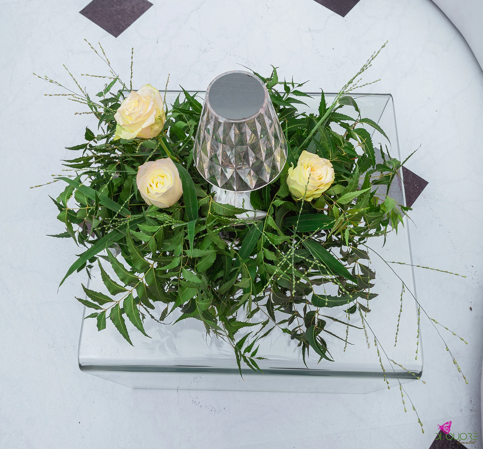 Mesa baixa de vidro com uma coroa de folhagem, três rosas claras e um candeeiro facetado, sobre o chão axadrezado a preto e branco