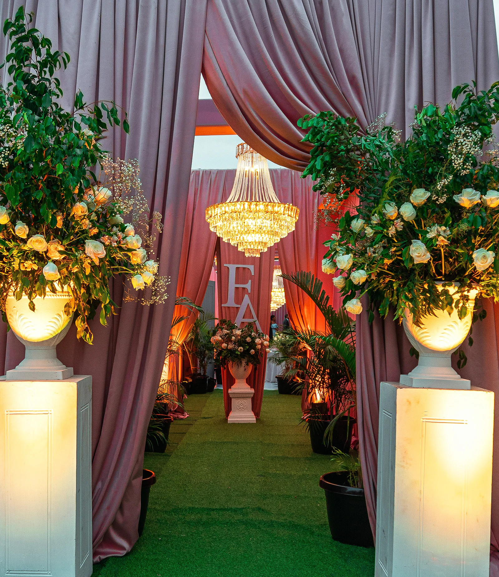 Entrada ao entardecer entre duas urnas iluminadas com rosas brancas sobre pedestais, cortinados cor de malva e, ao fundo, as iniciais F e A sob um lustre de cristal