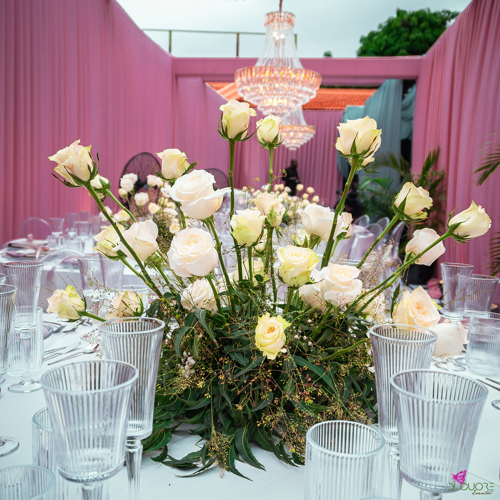 Arranjo de rosas claras entre copos canelados numa mesa posta, com lustres de cristal e cortinados cor-de-rosa ao fundo